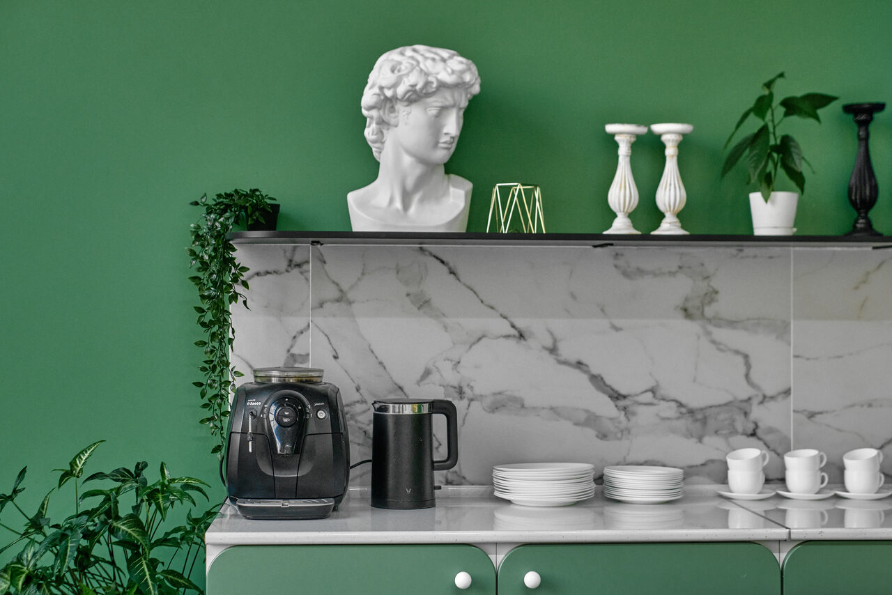 Арендовать Arcobaleno Loft(Аркобалено Лофт). Кухонная зона с мраморной столешницей и задней панелью. На столе кофеварка, чайник, стопка тарелок и чашек. На полке статуэтка головы, подсвечники, декор и растения. Стены зелёного цвета.