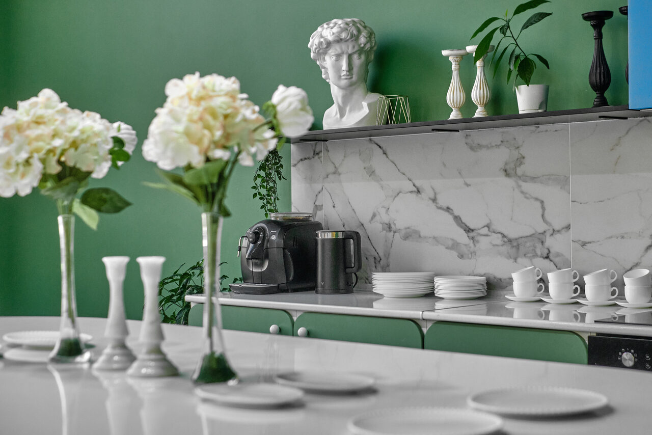 Арендовать Arcobaleno Loft(Аркобалено Лофт). Современная кухня с белым столом, зелеными шкафами и мраморной стеной. Белые тарелки и чашки на полке. Свежие цветы в вазах. На фоне бюст, вазы, кофеварка, растения. Настенное освещение.