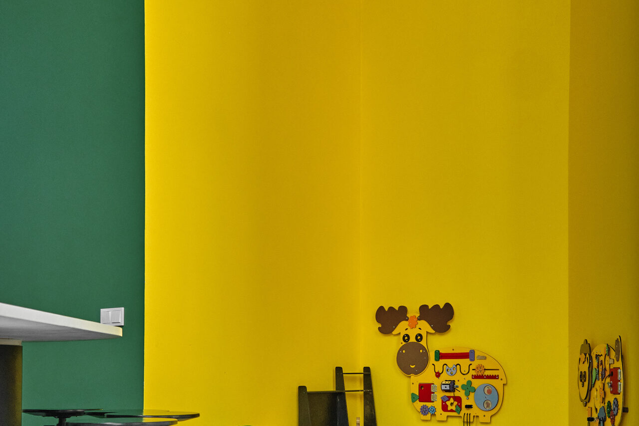 Арендовать Arcobaleno Loft(Аркобалено Лофт). Яркое помещение с желтыми и зелеными стенами. В углу на стене находится настенная игрушка в виде лося с элементами для игры. Видна часть белой столешницы. Освещение естественное.