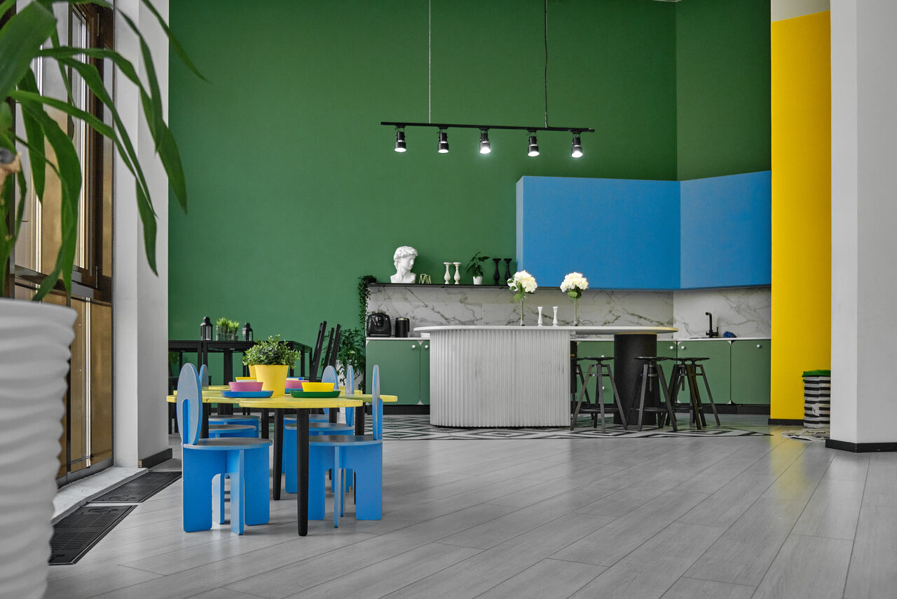 Арендовать Arcobaleno Loft(Аркобалено Лофт). Просторная кухня с зелёными и жёлтыми стенами, в центре круглый белый стол с тремя чёрными барными стульями, синие детские стулья и стол, подвесное точечное освещение, белый мраморный фартук, зелёно-голубые шкафы, большие окна, растения в вазонах.