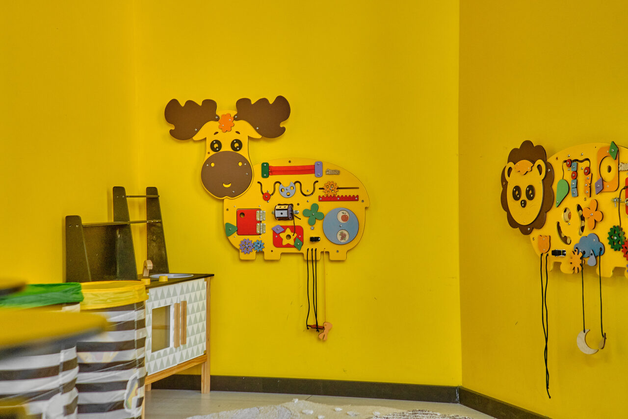Арендовать Arcobaleno Loft(Аркобалено Лофт). Детская комната с ярко-жёлтыми стенами, настенные панели в виде животных с развивающими элементами, деревянный стол с белыми и жёлтыми деталями, пластиковые корзины. Пол светлый, однотонный.