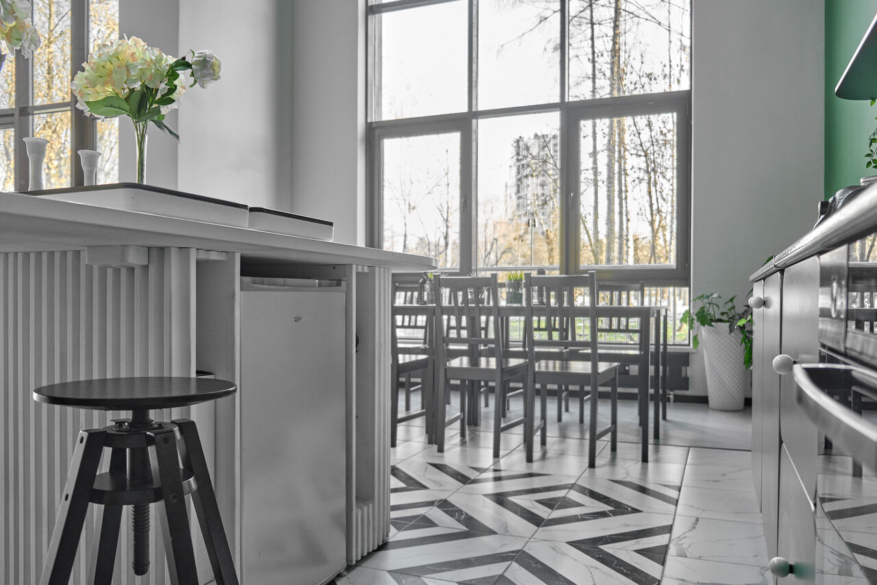 Арендовать Arcobaleno Loft(Аркобалено Лофт). Светлое помещение с большими окнами, столами и стульями из тёмного дерева, барная стойка с белым ребристым фасадом и табуретом. На стойке вазы с цветами. Пол с геометрическим узором, светло-серые тона.