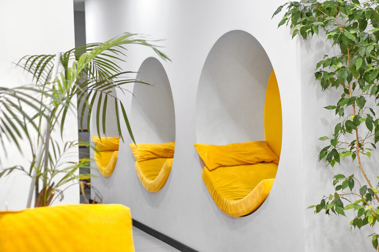 Арендовать Arcobaleno Loft(Аркобалено Лофт). Современное пространство с белыми стенами и круглыми нишами, содержащими желтые мягкие подушки. Растительность в горшках, контрастные элементы дизайна. Освещение мягкое, равномерное.