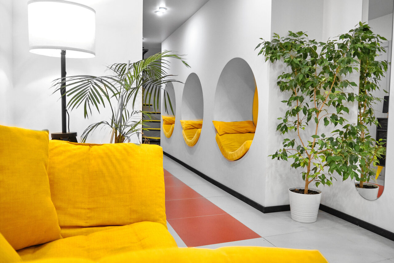Арендовать Arcobaleno Loft(Аркобалено Лофт). Современное пространство с белыми стенами и плиточным полом. Левая часть кадра: желтый диван и лампа. Справа: круглые ниши в стене с желтыми подушками. Растения в горшках, яркий свет.