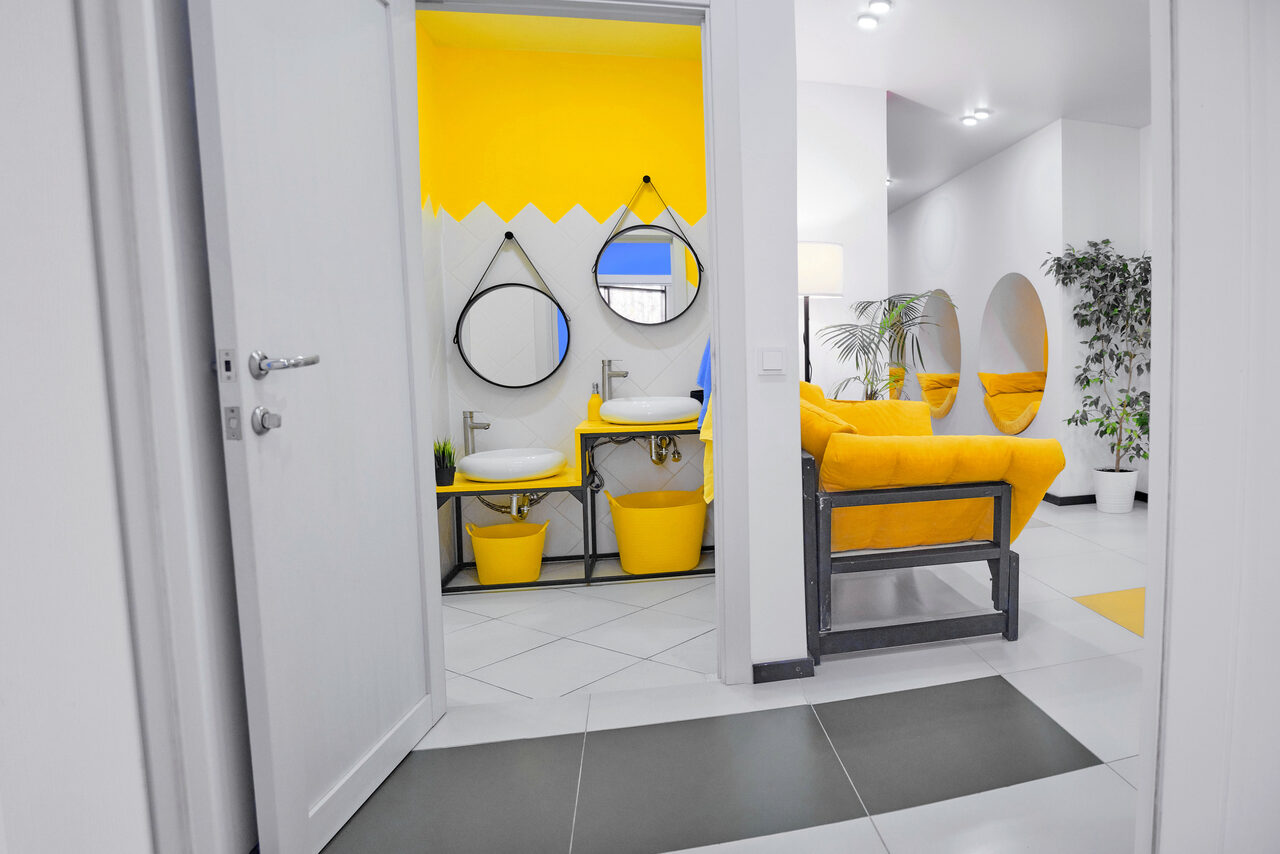 Арендовать Arcobaleno Loft(Аркобалено Лофт). Современное пространство с ярким дизайном. Зона умывальников с зеркалами и желтыми элементами. В комнате желтые диваны, растения и круглое зеркальное окно. Светлые стены, плиточный пол в серо-белых тонах, сочетание прямых и округлых форм.