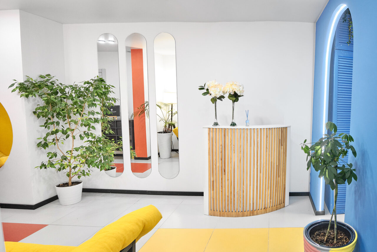 Арендовать Arcobaleno Loft(Аркобалено Лофт). Современное пространство с белыми стенами и одной синей, округлая стойка с деревянными планками, желтые диваны, растения в горшках, встроенные зеркала, яркое освещение, полы светло-серого цвета, нет текстов или знаков.