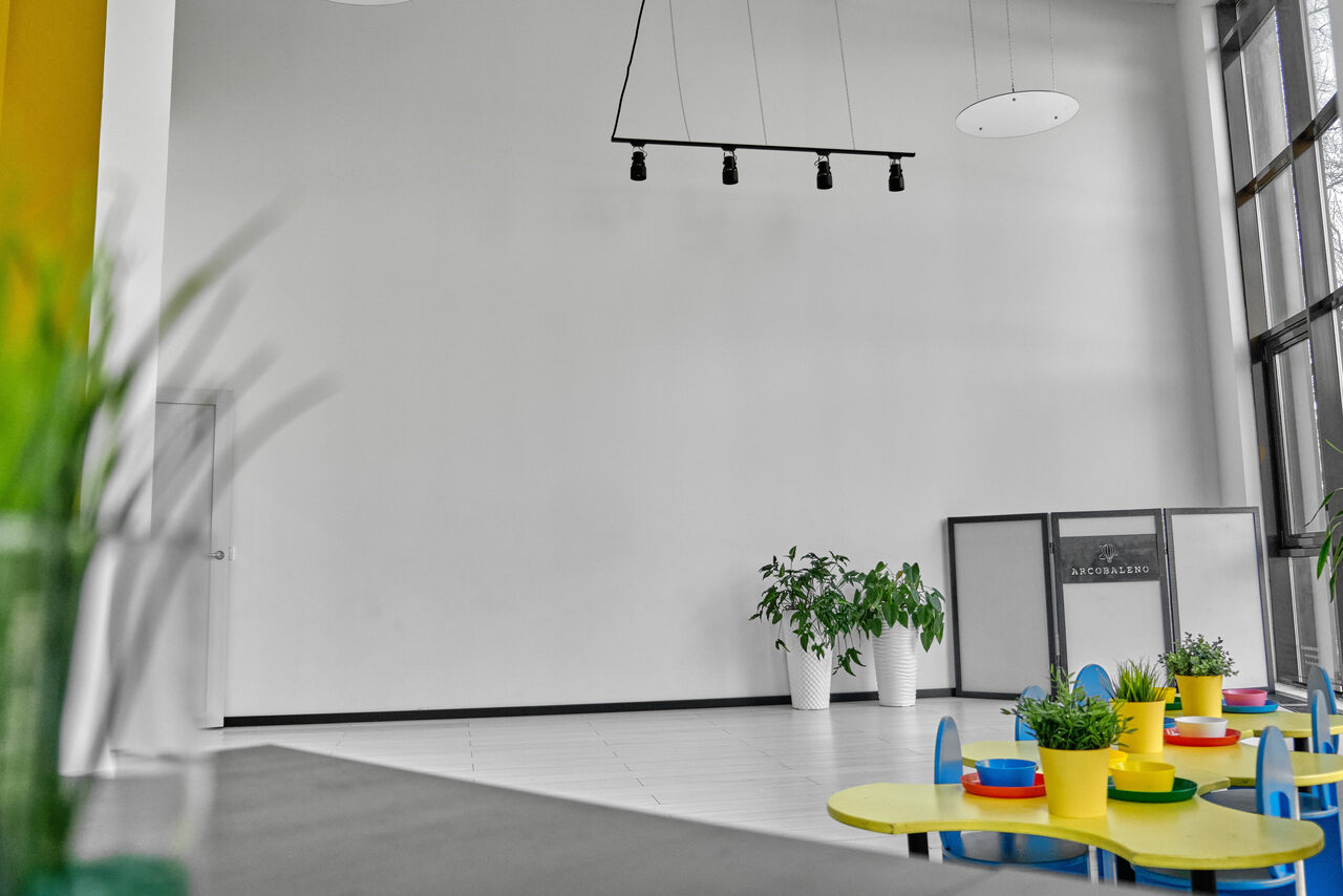 Арендовать Arcobaleno Loft(Аркобалено Лофт). Просторное светлое помещение с высоким потолком, белыми стенами и большими окнами. На переднем плане детские круглые столы с цветными горшками для растений. Слева зелёные растения, справа стеклянная перегородка. Потолочное освещение.