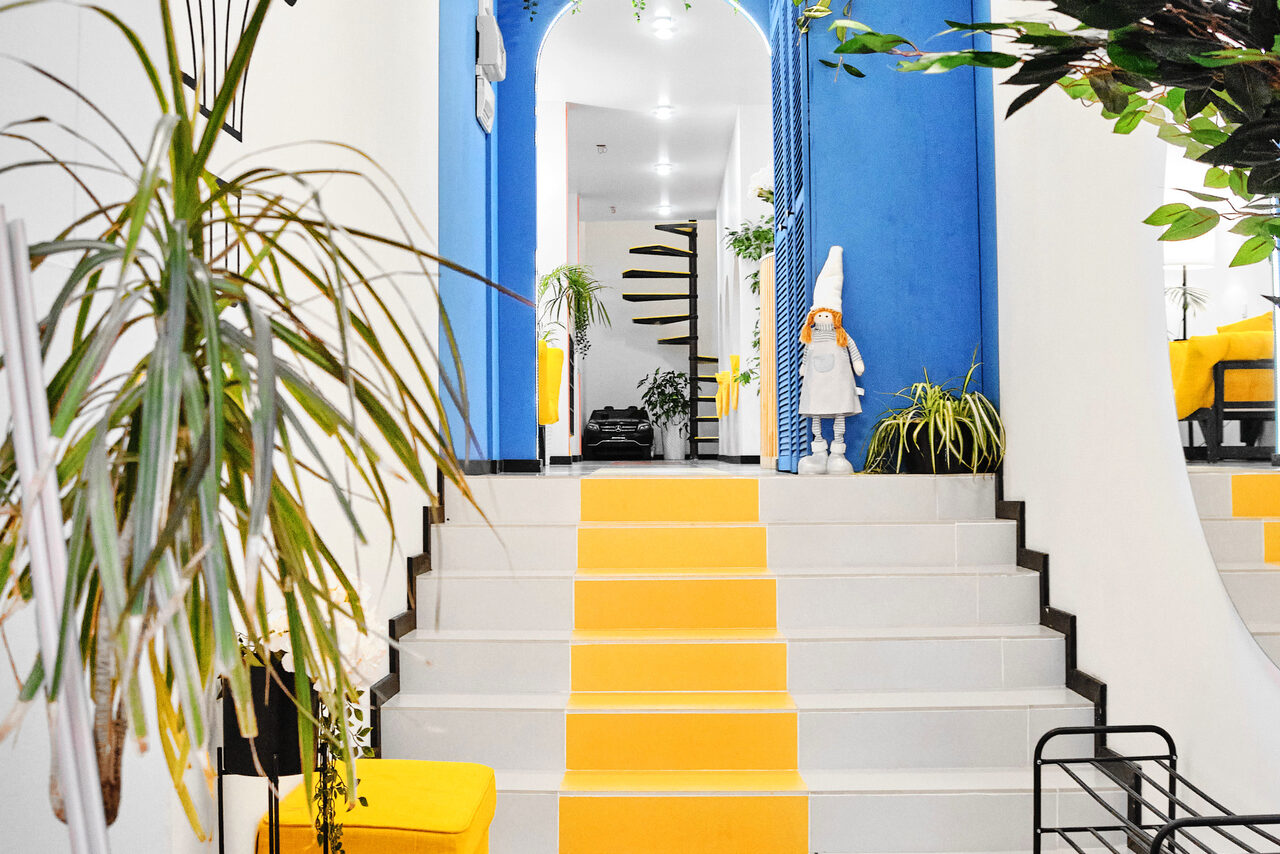 Арендовать Arcobaleno Loft(Аркобалено Лофт). Современное пространство с яркими синими и белыми стенами. Лестница с серыми и жёлтыми ступенями. Растения в горшках, жёлтый стул, чёрный металл стеллаж. Арочная дверь ведёт в комнату с чёрной спиральной лестницей. Акцентные освещённые зоны.