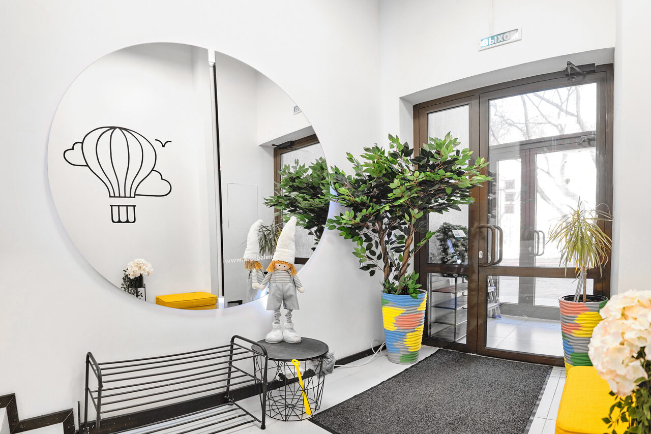 Арендовать Arcobaleno Loft(Аркобалено Лофт). Пространство с круглым зеркалом на стене, рисунком воздушного шара, рядом металлическая скамья, искусственные растения в цветных кашпо, статуя в шляпе, стеклянные двери, белые стены, декоративные элементы в светлых и жёлтых тонах.