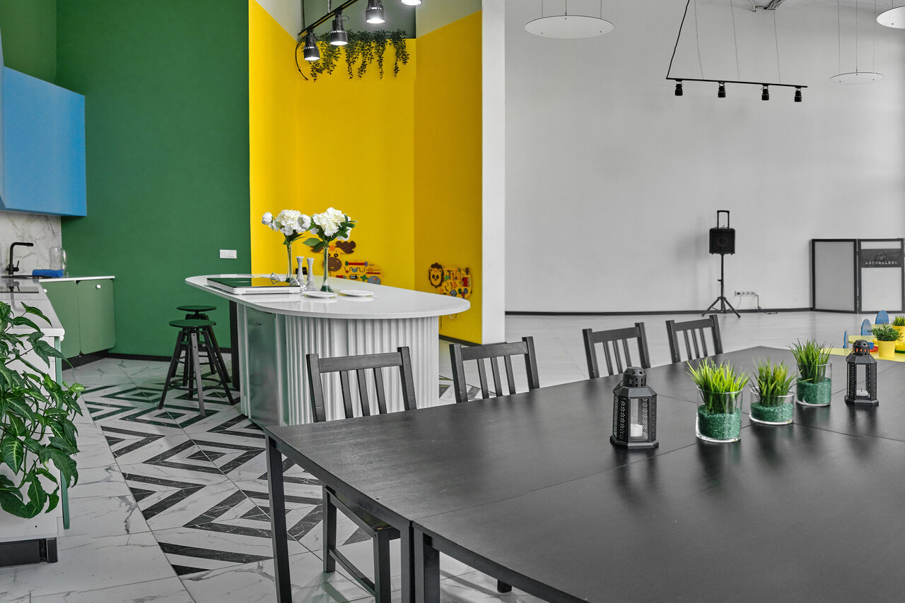 Арендовать Arcobaleno Loft(Аркобалено Лофт). Многофункциональное пространство с кухонной зоной и зоной для встреч. Белые и черные столы, черные стулья. Барная стойка с высокими стульями. Стены зеленые, желтые и белые. Растения и светильники. Пол с черно-белыми геометрическими узорами.