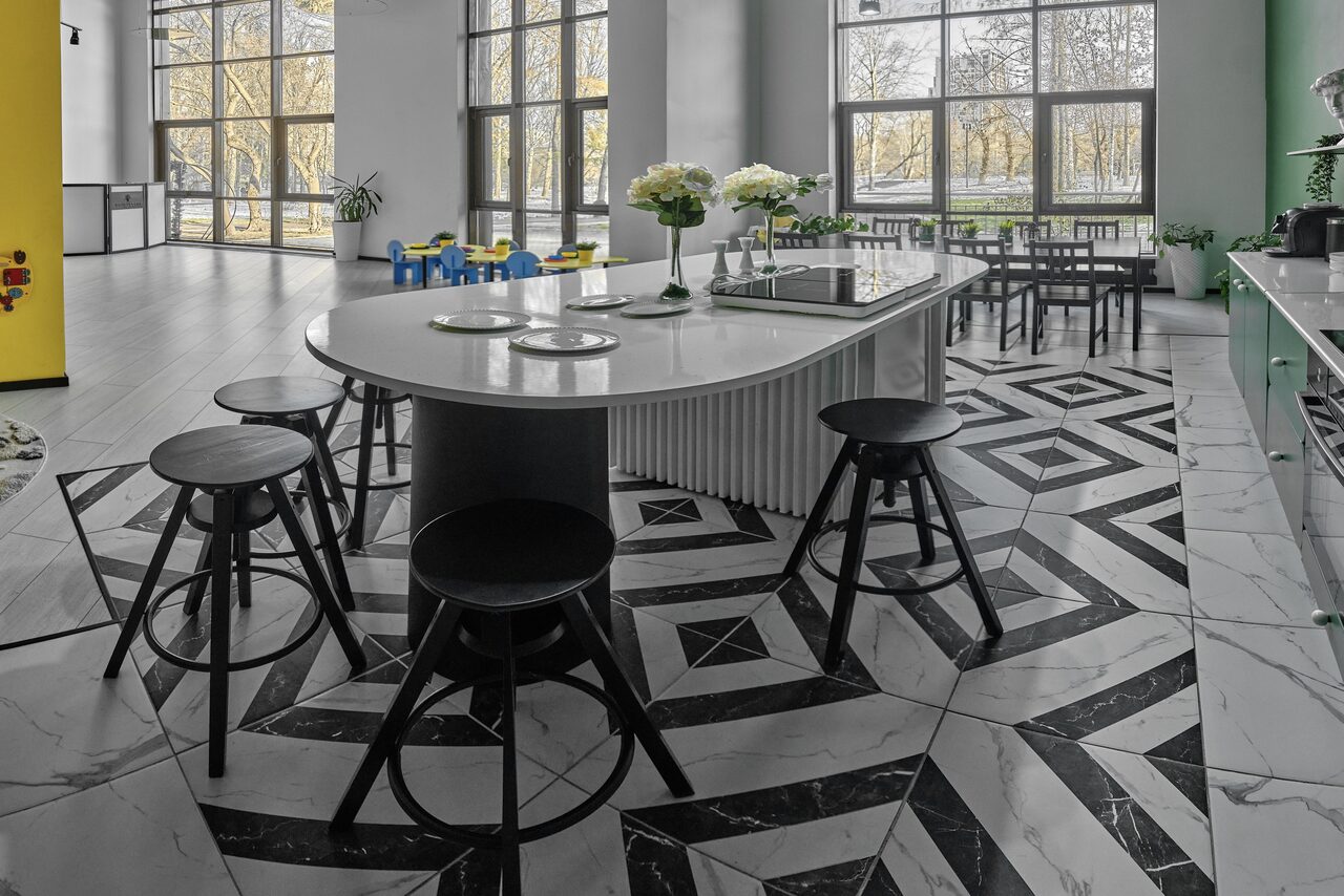 Арендовать Arcobaleno Loft(Аркобалено Лофт). Современное просторное помещение с большими окнами и центральным островом на плиточном полу. Белый стол окружен черными стульями. В комнате есть зелёная стена, кухня с бытовой техникой и детская зона с яркими стульями и столиками.