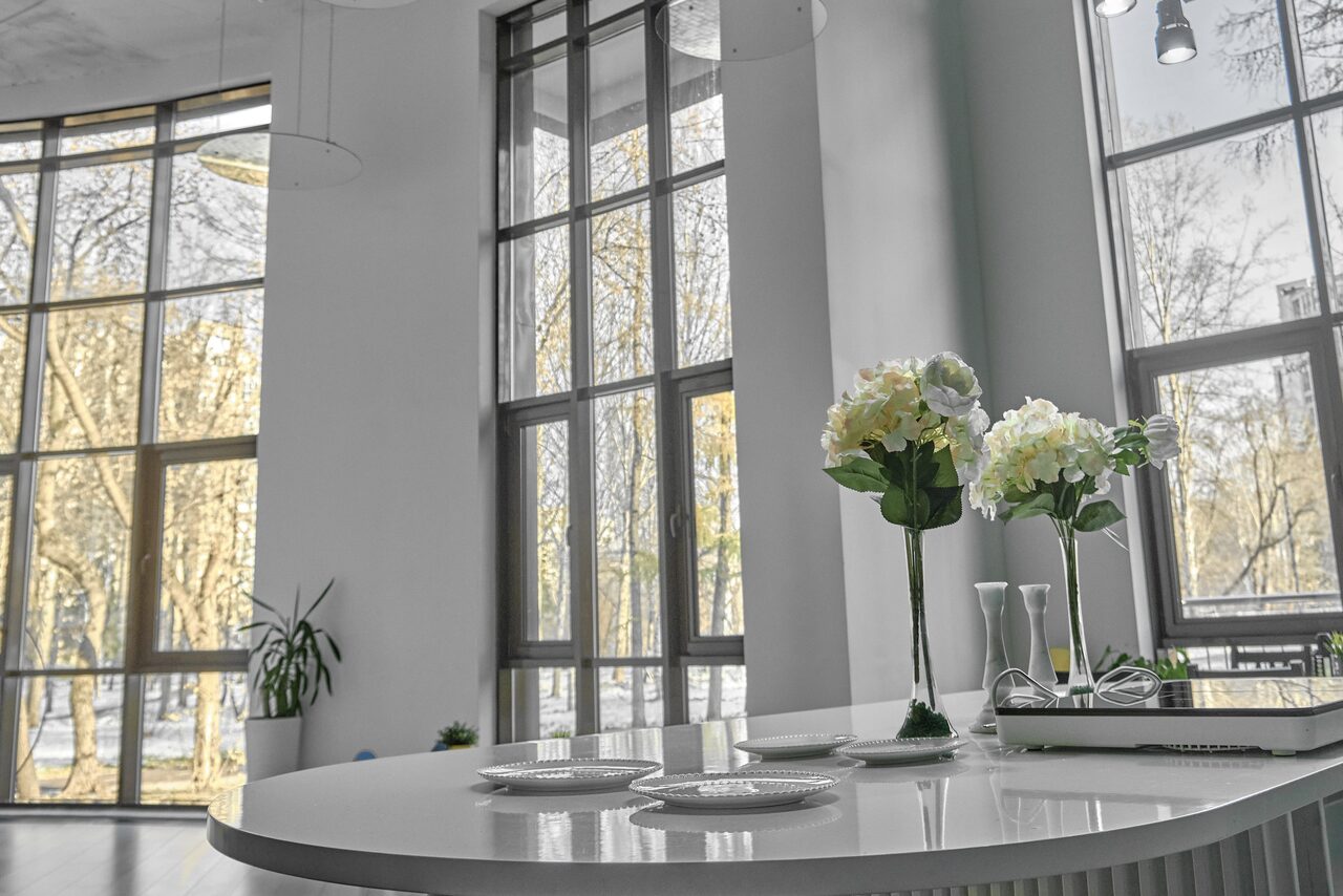 Арендовать Arcobaleno Loft(Аркобалено Лофт). Современное светлое помещение с большими окнами, овальным белым столом, цветочными декоративными элементами и тарелками. Светлые стены, подвесные светильники и вид на улицу с деревьями за окнами.