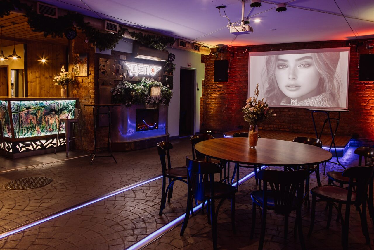 Арендовать Depo Loft(Депо Лофт). Пространство представляет собой уютный зал с кирпичными стенами. Центр занимает круглый деревянный стол с черными стульями. Слева барная стойка с подсветкой, справа проектор с изображением. Потолочный свет, неоновая подсветка пола.