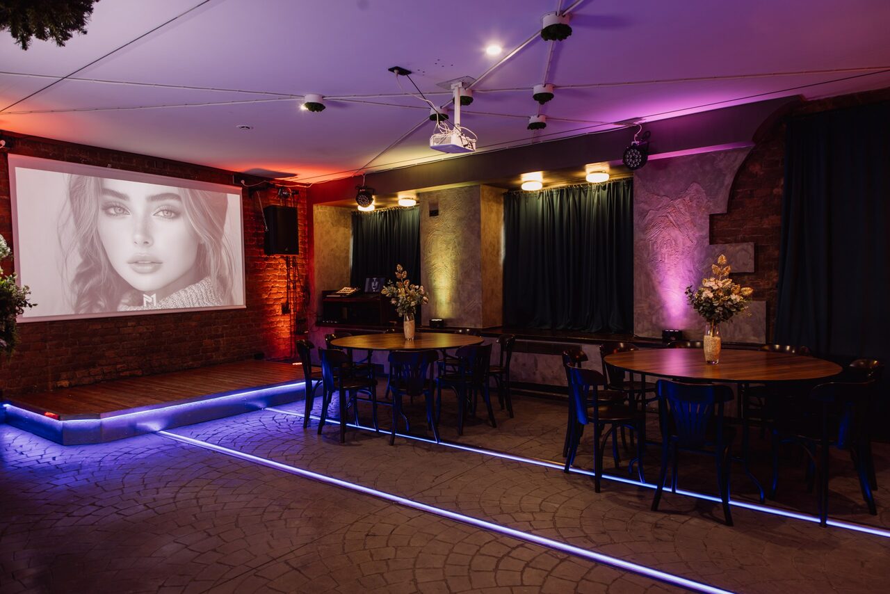 Арендовать Depo Loft(Депо Лофт). Просторное помещение для мероприятий с кирпичными стенами и крупным экраном для проекций. На экране изображение лица. Несколько круглых столов с черными стульями. Яркое разноцветное освещение, драпировки на стенах, декор из цветов.