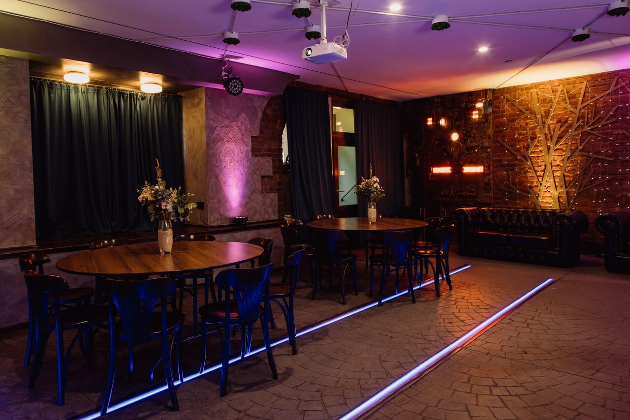Арендовать Depo Loft(Депо Лофт). Интерьерное пространство с двумя круглыми деревянными столами, окружёнными черными стульями. На каждом столе вазоны с цветами. Темные шторы, кирпичная стена с декоративной подсветкой, мягкий кожаный диван. Натяжной потолок с лампами, проектор и светодиодные линии на полу.