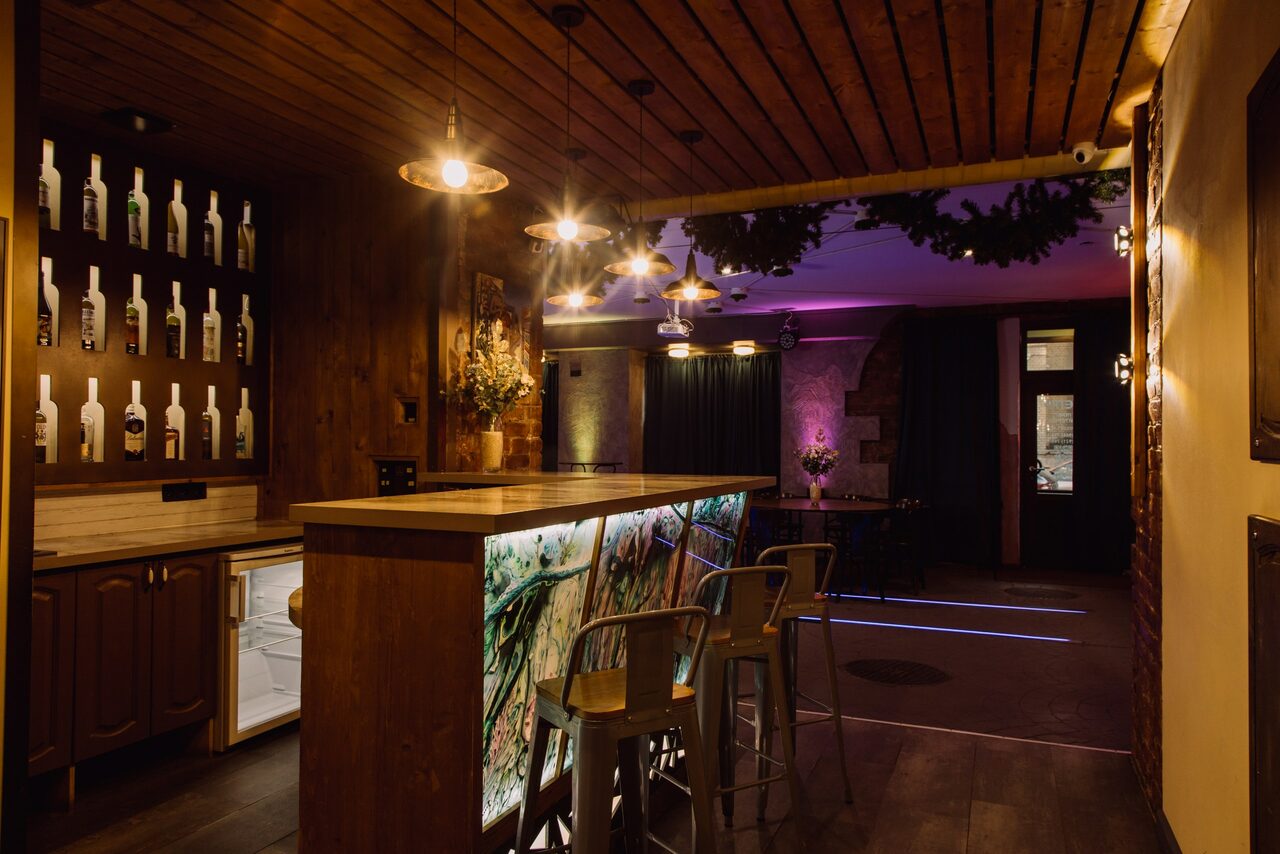 Арендовать Depo Loft(Депо Лофт). Коктейльный бар с деревянными стенами и потолком, светящаяся барная стойка с высоким стульями, стена с полками для бутылок, подвесные светильники, стол из темного металла и темная занавеска в интерьере, мягкое разноцветное освещение.