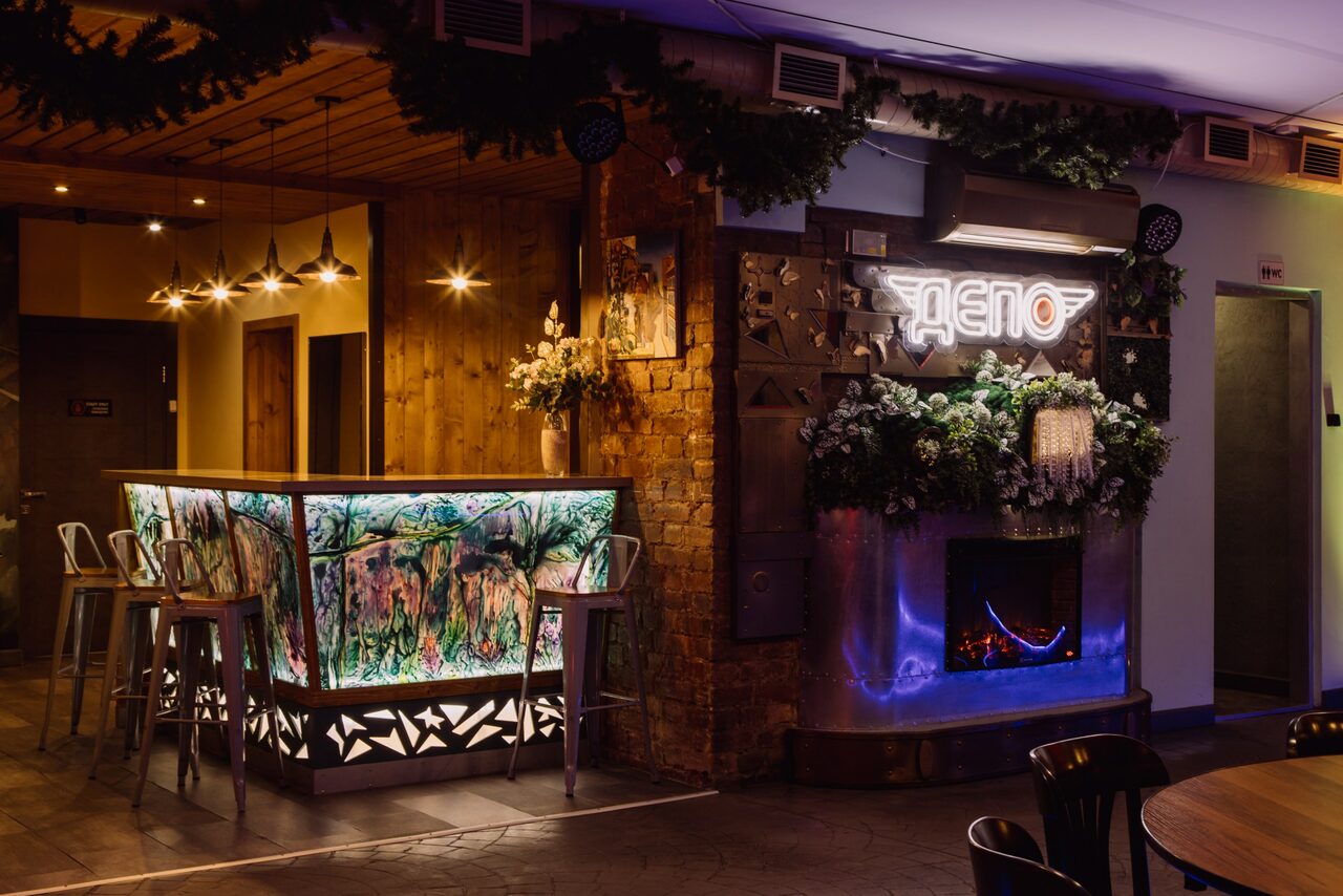 Арендовать Depo Loft(Депо Лофт). Барная зона с декоративной стойкой, украшенной разноцветными изображениями, высокие металлические стулья. Справа неоновая вывеска «ДЕПО», стилизованный камин. Декор из зелени и цветов. Потолок деревянный, светильники подвесные. Тёмные тона интерьера.