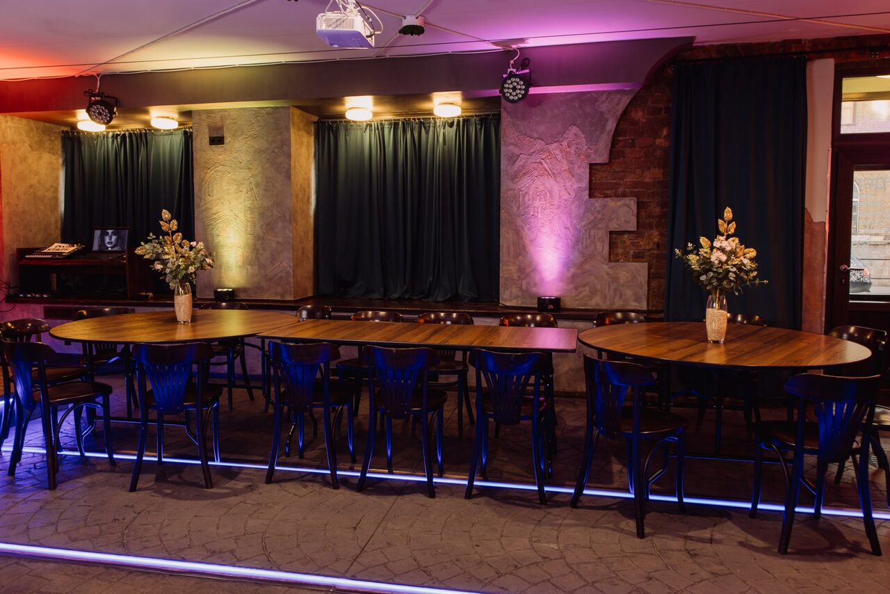 Арендовать Depo Loft(Депо Лофт). Пространство с длинным деревянным столом и черными стульями. Декорировано вазами с цветами. Освещение включает настенные светильники и подвесные прожекторы. Стены из кирпича и штукатурки, тёмные шторы. Пол с плиткой, подсветка вдоль пола.