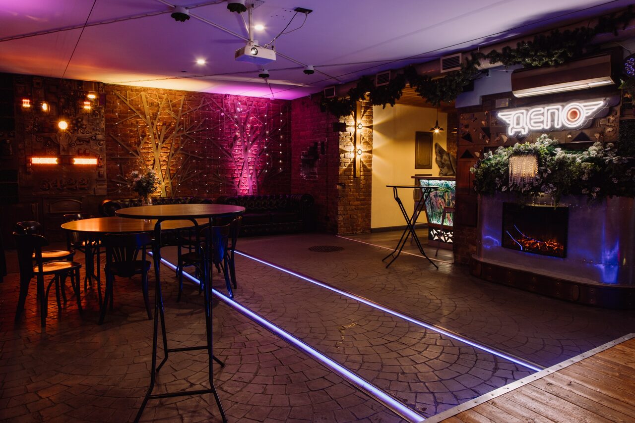 Арендовать Depo Loft(Депо Лофт). Пространство с декоративной кирпичной стеной и неоновым знаком, круглый деревянный стол и стулья, барная стойка с растениями, мягкое разноцветное освещение, светодиодные полосы на полу, проекционное оборудование на потолке.