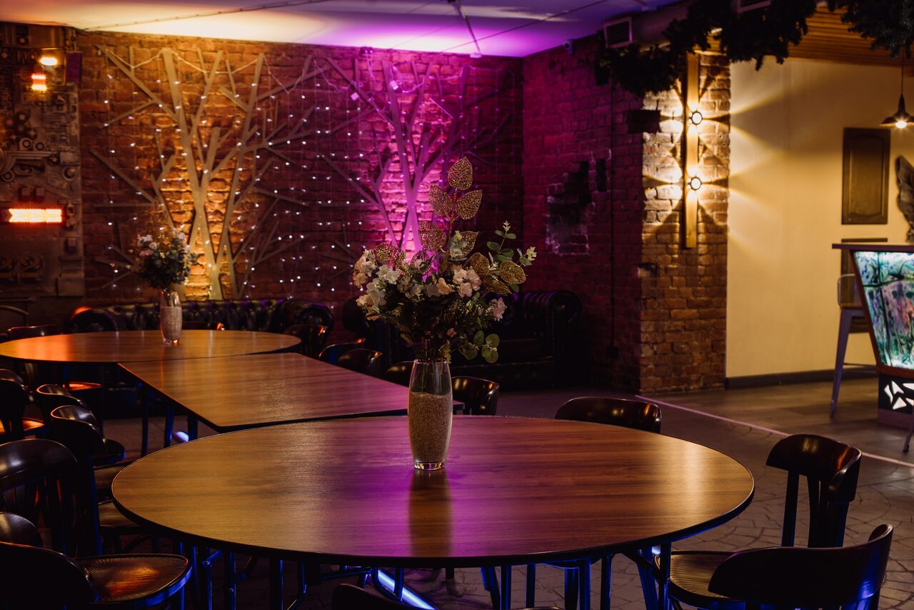 Арендовать Depo Loft(Депо Лофт). Овальный деревянный стол с вазой цветов в помещении с кирпичными стенами. На стене узор из светодиодов в форме деревьев. Мягкое разноцветное освещение, декор зеленью, несколько ламп и консольных светильников.