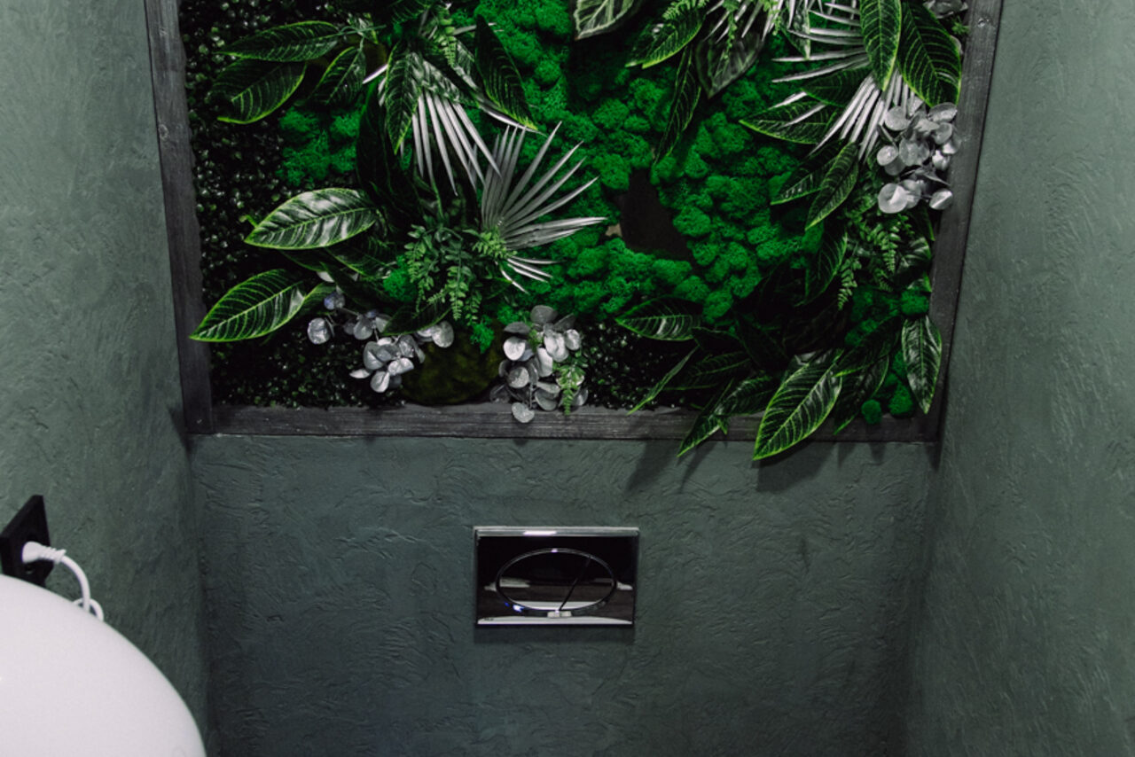 Арендовать Depo Loft(Депо Лофт). Зелёная стена с искусственными растениями, металлическая кнопка смыва, текстурированные серые стены, белый керамический унитаз частично виден слева.