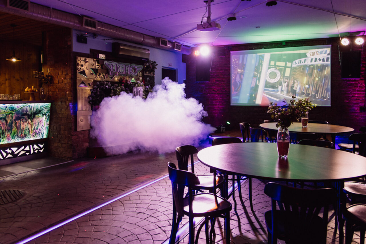 Арендовать Depo Loft(Депо Лофт). Зал с круглыми столами и стульями, кирпичная стена, проектор с изображением, дымовая машина, разноцветное освещение. На столах цветочные композиции. Барная стойка с подсветкой. Потолочные вентиляторы и элементы декора.