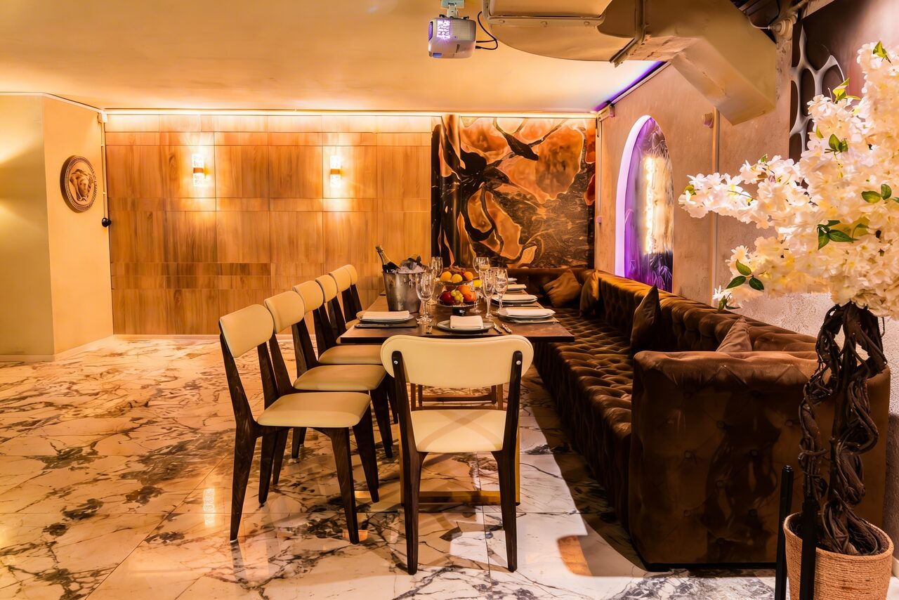 Арендовать Стильный лофт Джаванди. Элегантное пространство с большим прямоугольным столом, обставленным стульями и мягким диваном. Мраморный пол, потолочные светильники, деревянная стена с подсветкой. Украшение из белых цветов и настенное искусство. Светлая и уютная обстановка.