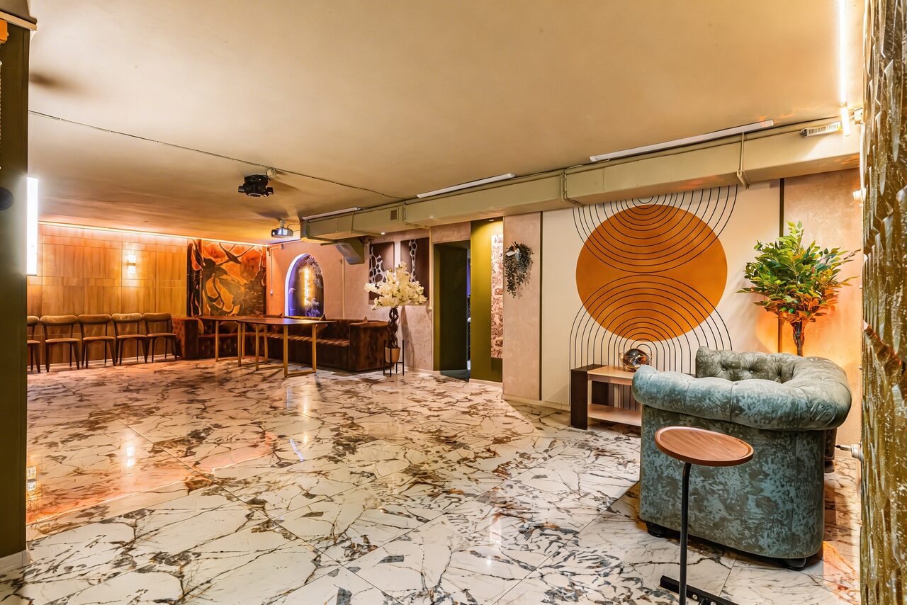 Арендовать Стильный лофт Джаванди. Просторное помещение с мраморным полом. В центре — изогнутый диван и круглый столик. Стены украшены настенной графикой с кругами. Барная стойка и высокие стулья у стены. Потолочное освещение и проектор. Декоративные растения в вазах.