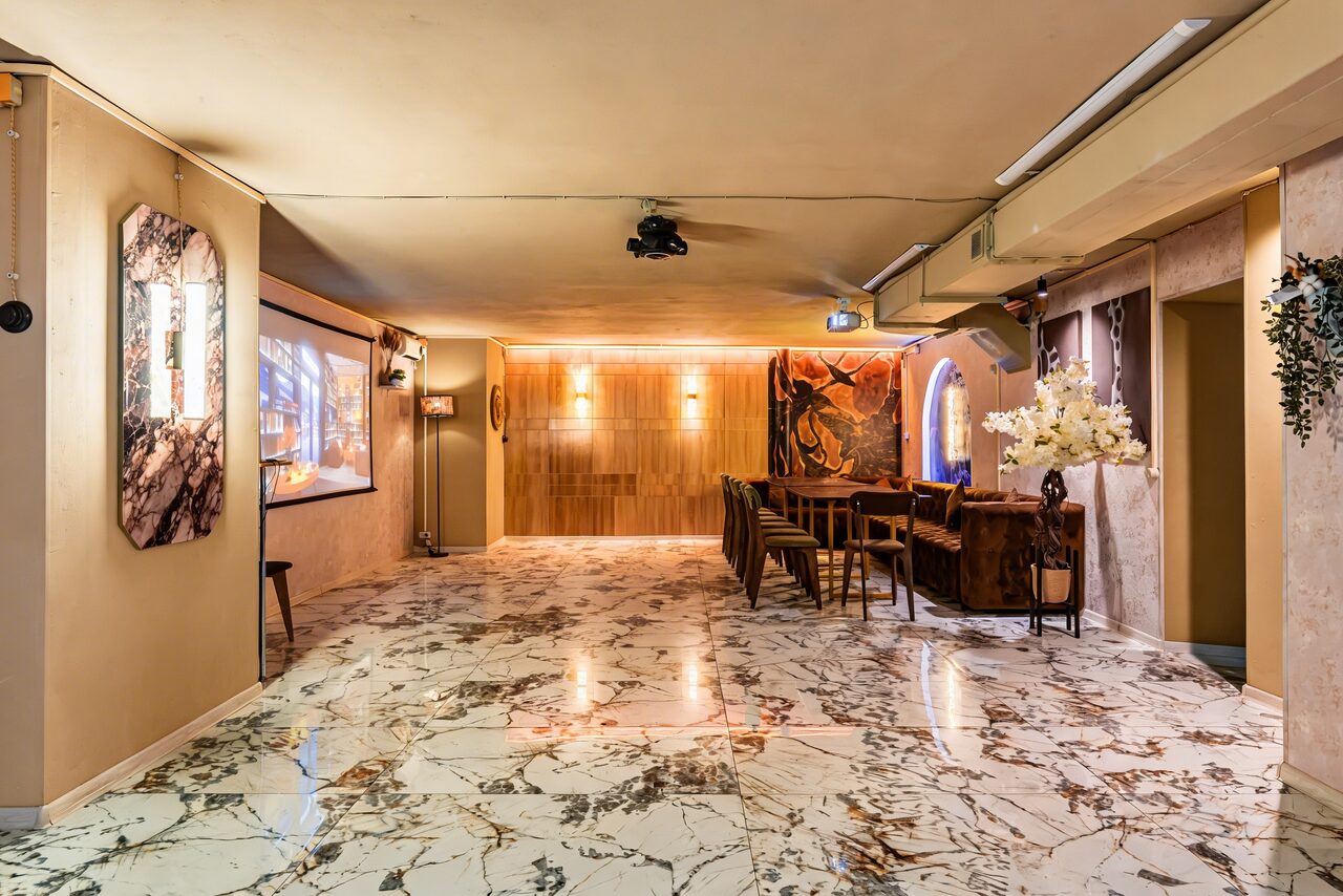 Арендовать Стильный лофт Джаванди. Мраморный пол с разноцветными прожилками, нейтральные стены, деревянная панель на дальней стене, несколько стульев и стойка, проектор на потолке, настенные картины и декор, мягкое освещение, крупная ваза с белыми цветами.