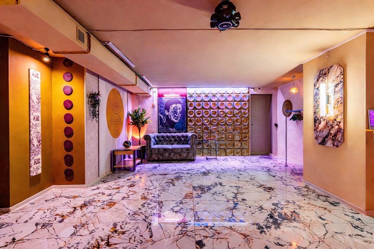 Арендовать Стильный лофт Джаванди. Пространство с мраморным полом, кожаным диваном у стены с декоративными тарелками и неоновой подсветкой. Потолок светлый, светильники на стенах и потолке. Колонны украшены панелями и декоративными элементами. Стены светлых тонов.