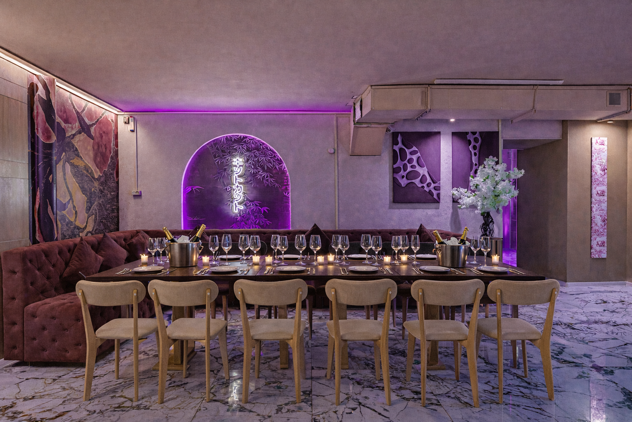 Арендовать Стильный лофт Джаванди. Большое помещение с длинным обеденным столом и деревянными стульями. Большой диван вдоль стены, где фиолетовая подсветка и декоративные панели. На столе стоят бокалы и металлические ведра. Мраморный пол, бежевые стены.