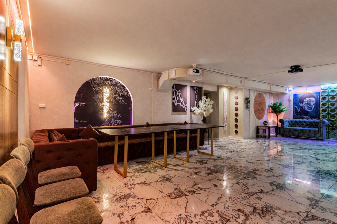 Арендовать Стильный лофт Джаванди. просторное помещение с мраморным полом, длинный деревянный стол с мягкими креслами, плюшевые диваны, настенные декорации с геометрическим узором и цветами, мягкое освещение, цветы в вазах, светло-бежевые стены, подвесная лампа, проектор на потолке