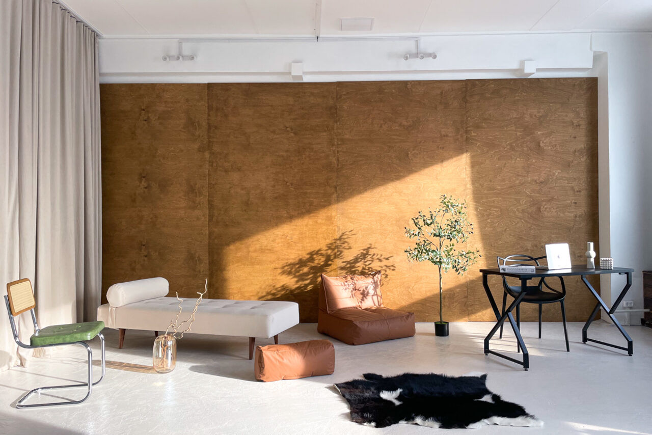 Арендовать Зал Серебро. Просторная и светлая фотостудия. Просторное помещение с белыми стенами и полом, акцентированное деревянной панелью. Мебель: белый диван, коричневые пуфы, черный стол, зеленый стул. Освещение естественное. Декор: растение в горшке, ковёр с животным принтом. Справа ноутбук на столе.