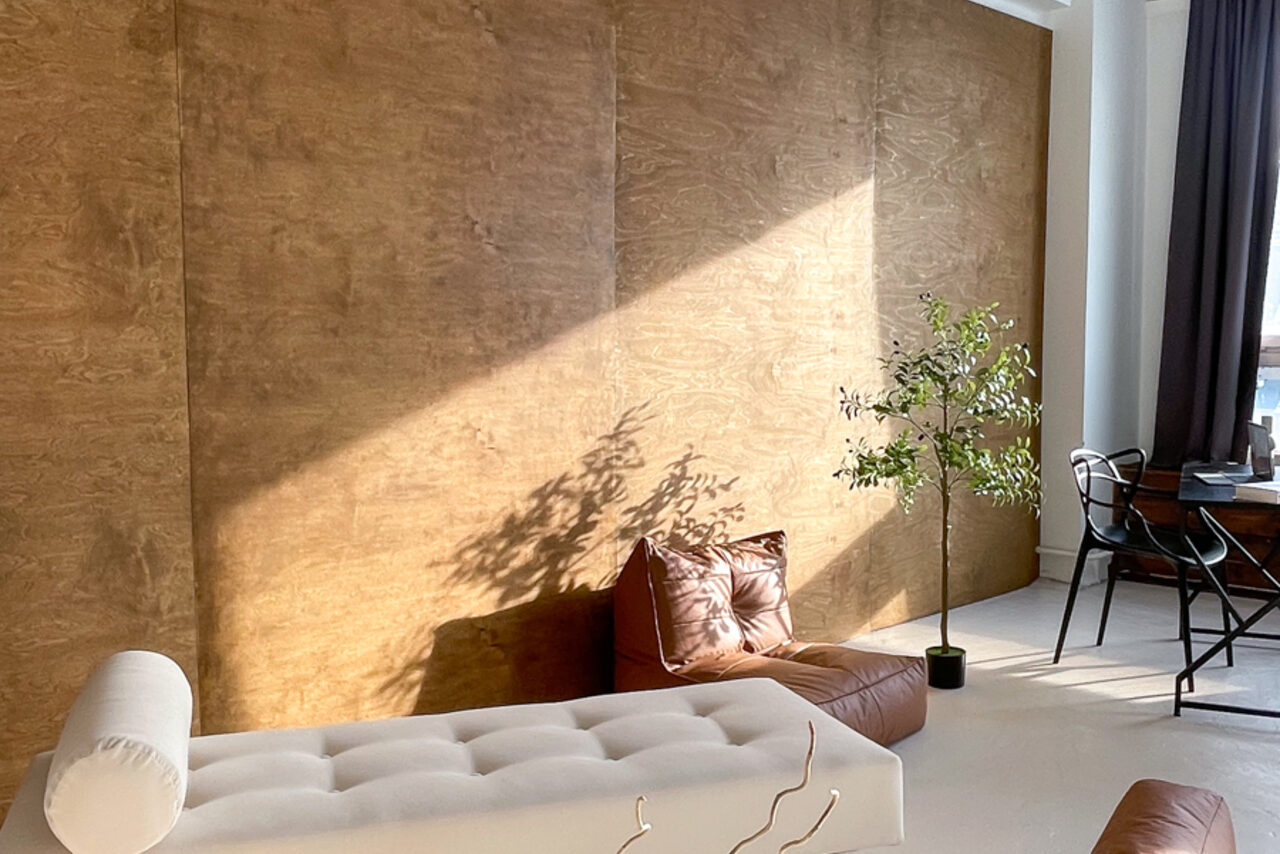 Арендовать Зал Серебро. Просторная и светлая фотостудия. Пространство с деревянной стеной, белым диваном и коричневой кожаной подушкой. У окна черный металлический стол с креслом, рядом зеленое растение в горшке. Естественное освещение, светло-серая штора, пол светлого оттенка.