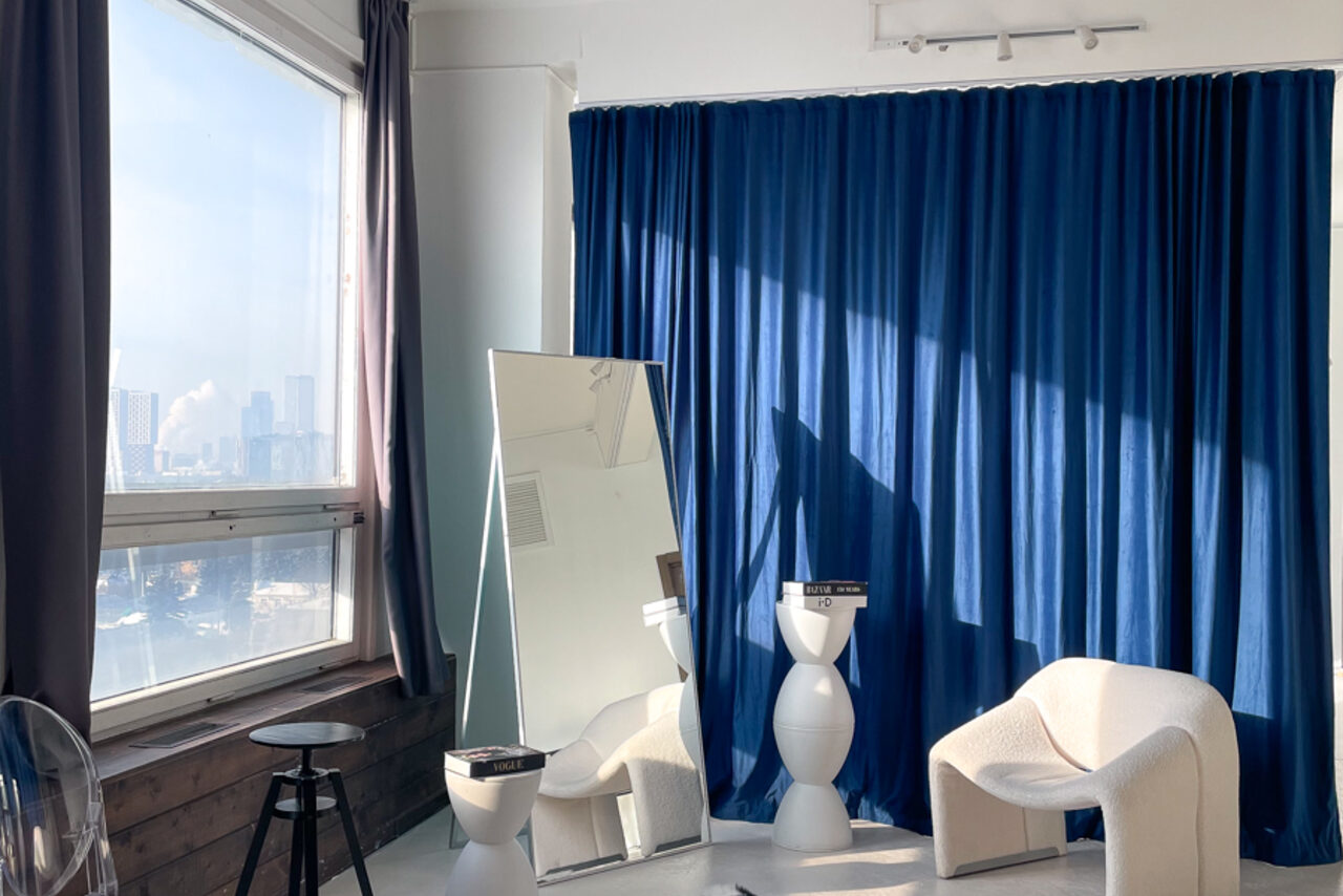 Арендовать Зал Серебро. Просторная и светлая фотостудия. Пространство со светлым креслом, высоким прямоугольным зеркалом, круглым белым столиком с книгами, тёмно-синей драпировкой на стене. Большое окно с серыми шторами, виден городской пейзаж. Слева деревянный стул, стены и пол светлые. Естественное освещение.