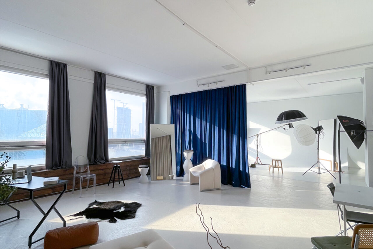 Арендовать Зал Серебро. Просторная и светлая фотостудия. Светлое фотостудийное пространство с большими окнами, серая штора, белые стены и пол, синий занавес, зеркала, стул, студийное осветительное оборудование, коврик с животным принтом, стол со стулом, мягкое кресло, деревянная скамья.