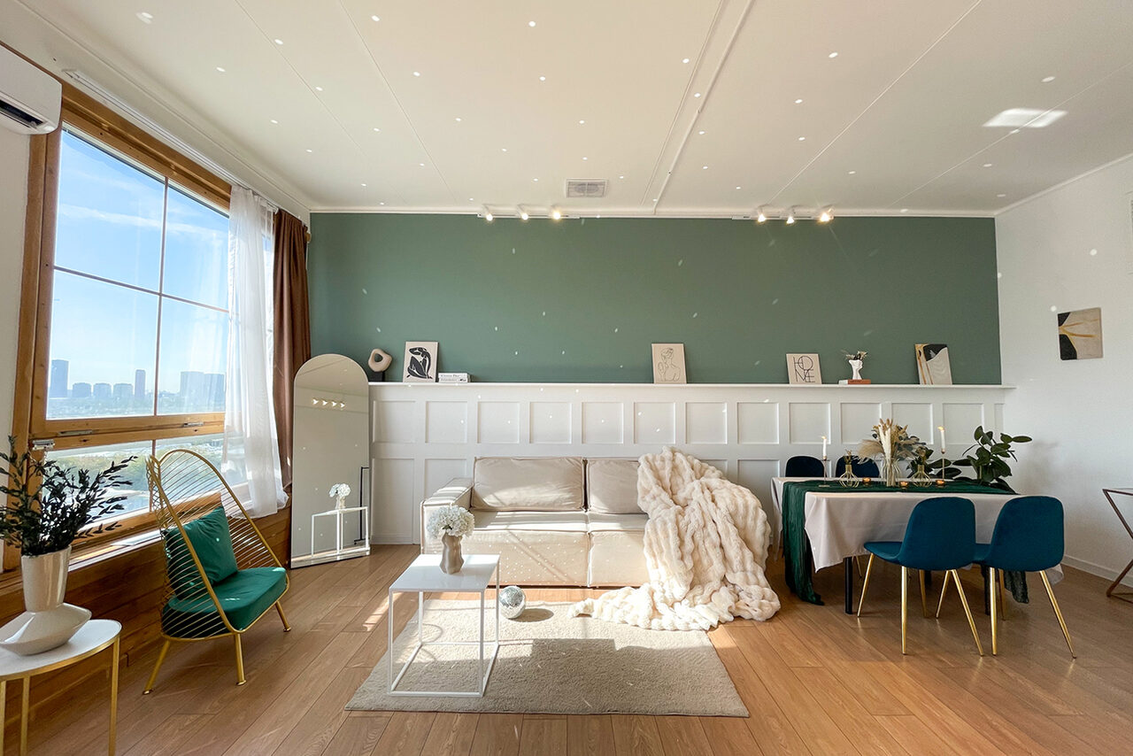 Арендовать Зал Квартира. Уютное пространство в квартирном стиле. Светлая комната с деревянным полом и зелёной акцентной стеной. Белый диван, зеркало в полный рост, небольшой столик, обеденный стол с синими стульями. Большое окно, шторы. Размещение предметов декора на полке. Подвесное освещение.