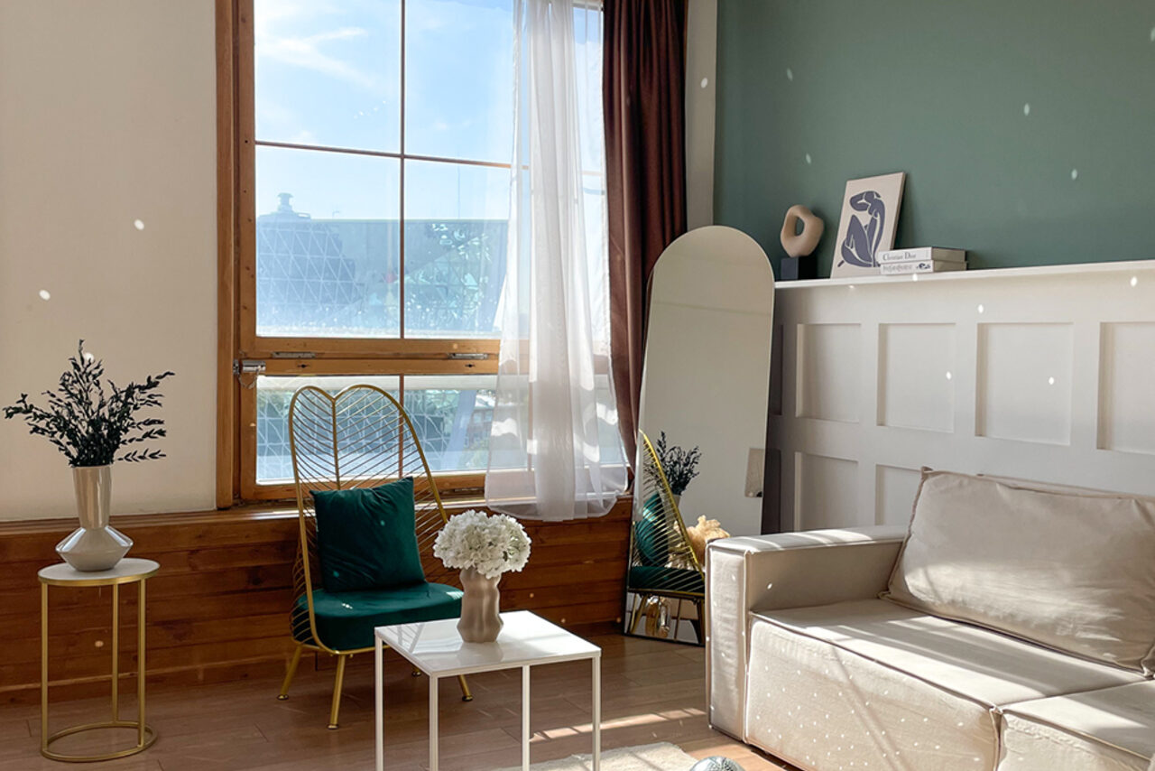 Арендовать Зал Квартира. Уютное пространство в квартирном стиле. Гостиная с большим окном, бело-зелеными стенами и деревянным полом. Мебель: бежевый диван, белый стол, золотистое кресло с зеленой подушкой. Декор: зеркало, вазы с цветами, книги. Естественное освещение, белые шторы, деревянные элементы.