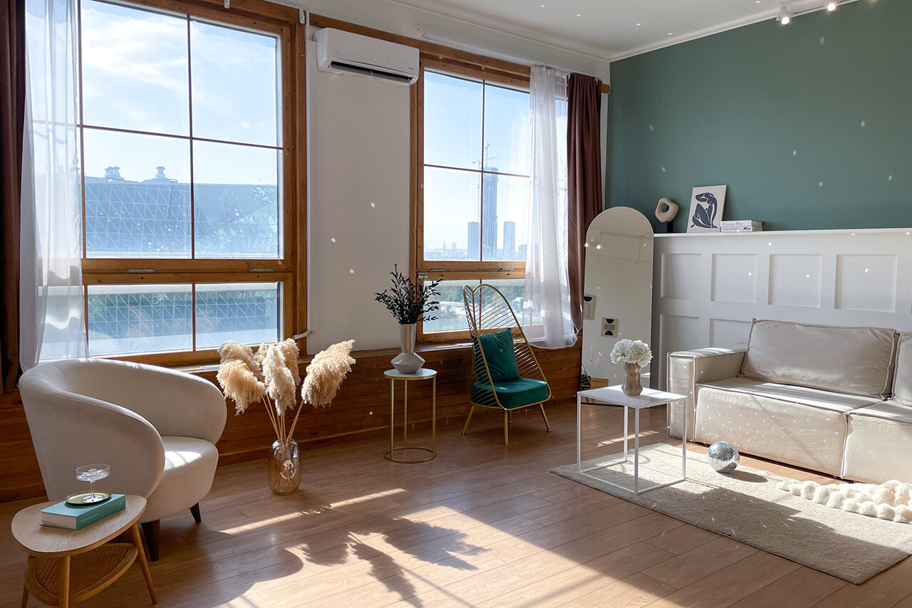 Арендовать Зал Квартира. Уютное пространство в квартирном стиле. Гостиная с деревянным полом, двумя большими окнами и бежевыми шторами. Светлый диван, два кресла, круглые столики. Стена зеленого цвета с деревянными панелями, есть декор в виде сухоцветов. Большое зеркало и кондиционер на стене. Естественное освещение.