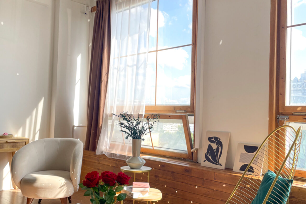 Арендовать Зал Квартира. Уютное пространство в квартирном стиле. Светлое помещение с большими окнами и длинными шторами, белое кресло, деревянный стол с букетом красных роз, декоративные картины на подоконнике, растение в вазе, деревянные элементы, светлый текстиль.