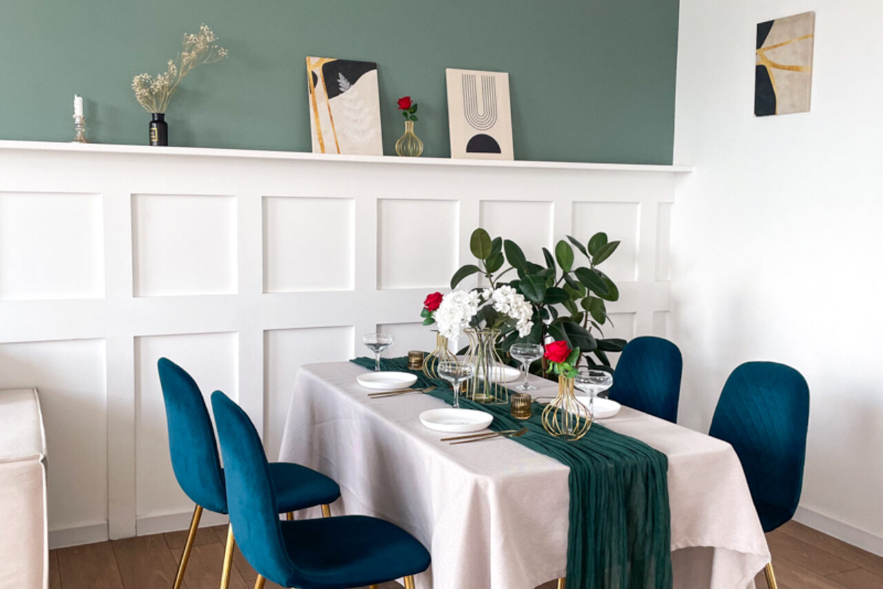 Арендовать Зал Квартира. Уютное пространство в квартирном стиле. Столовая зона с прямоугольным столом, накрытым белой скатертью и зелёной дорожкой. Синие стулья, белые панели на стене, зелёная стена сверху, картины и декор на полке. Светлое помещение с деревянным полом и декоративными растениями.