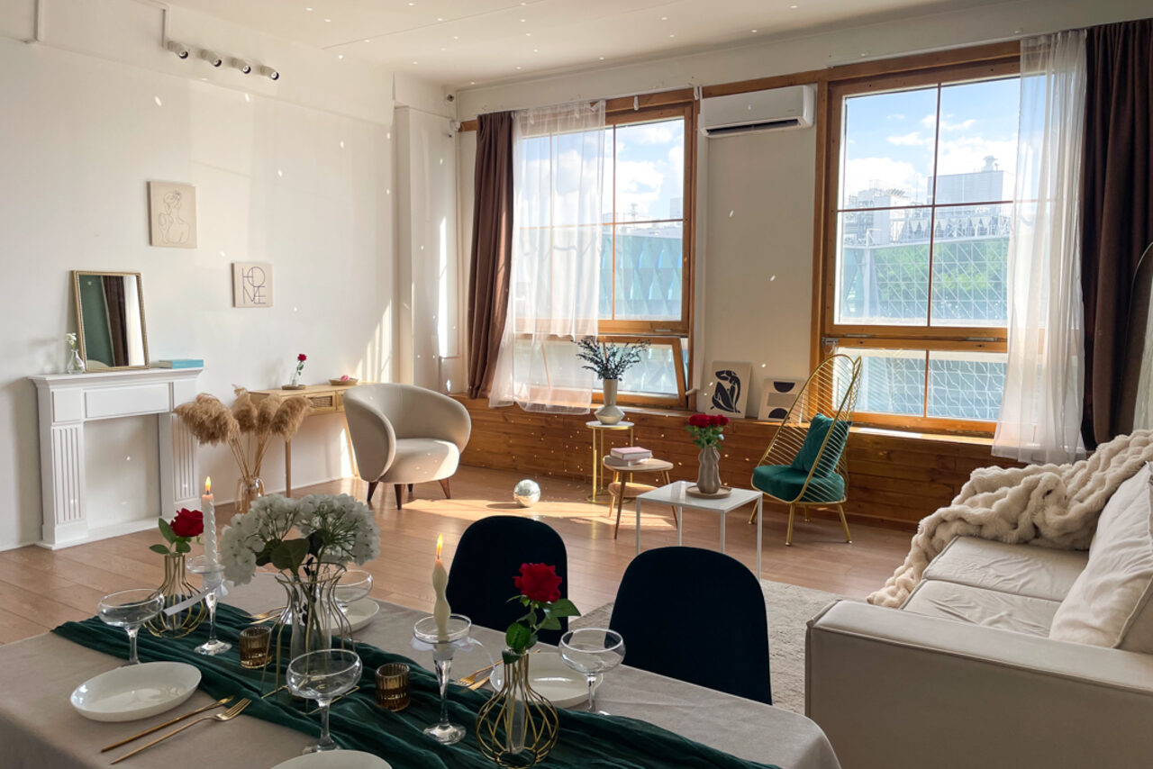 Арендовать Зал Квартира. Уютное пространство в квартирном стиле. Гостиная с обеденным столом, сервированным бокалами и цветами. Кресла и диван у больших окон с занавесками, декор: вазы, картины, зеркало. Стены белые, пол деревянный. Естественное светлое освещение, воздушные шары и растения в интерьере.