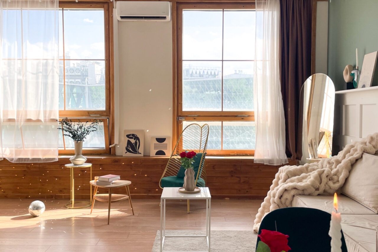 Арендовать Зал Квартира. Уютное пространство в квартирном стиле. Светлая комната с двумя большими окнами, занавешенными прозрачными шторами. Деревянный пол, светлая мебель: диван, кресло, два столика. На столах вазы с цветами и книгами. Зеркало в углу, кондиционер на стене, картины на подоконнике.