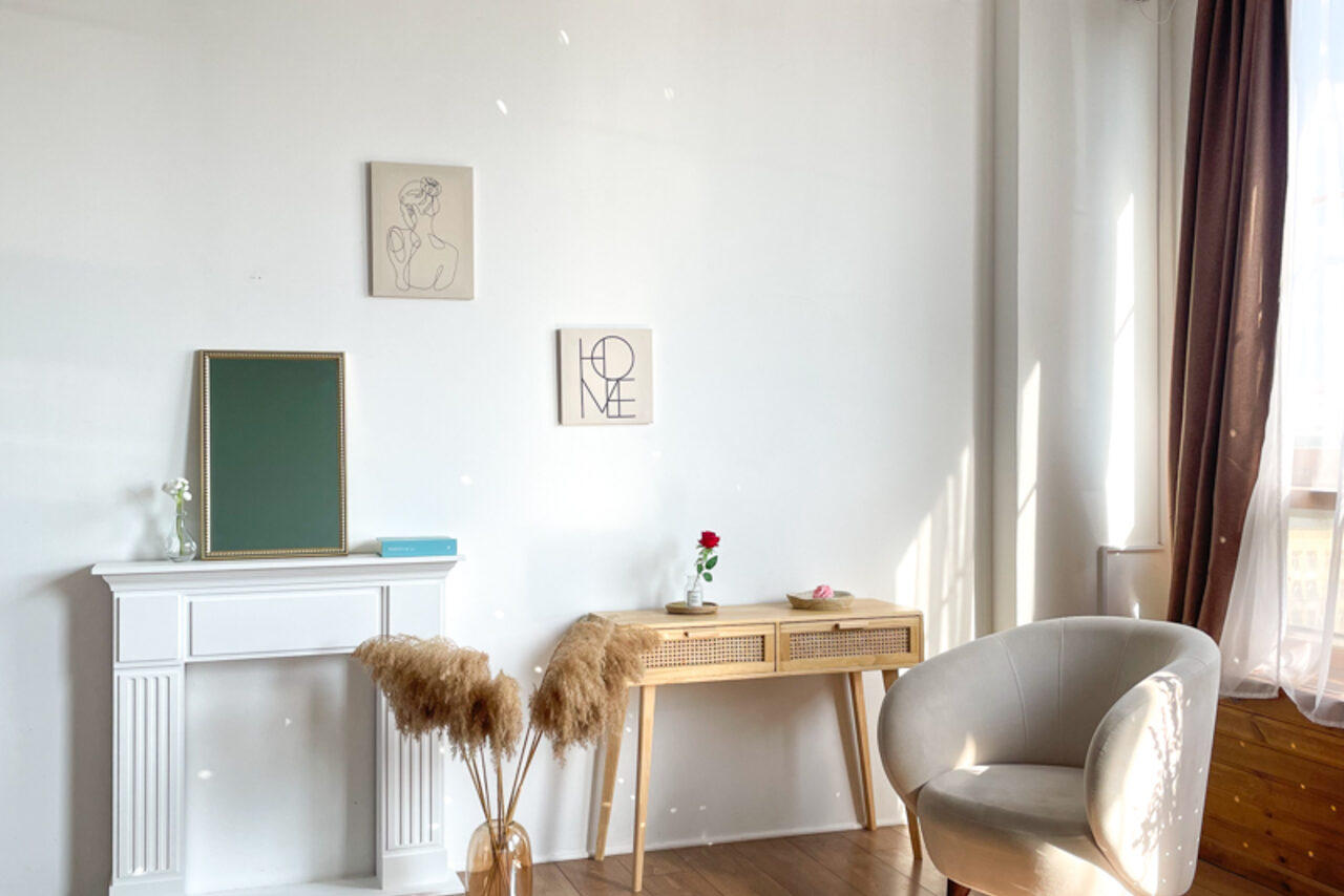 Арендовать Зал Квартира. Уютное пространство в квартирном стиле. Светлая комната с белыми стенами, деревянным полом. У стены белый камин, на нем зеленая доска и книга. Рядом вазон с сухоцветами. Деревянный стол, на нем роза в вазе. Кресло в уголке. На стенах два постера. Справа окно с прозрачной занавеской.