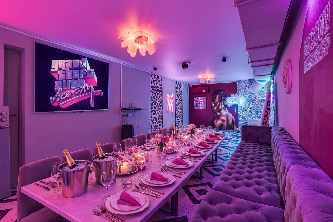 Арендовать Неоновый лофт Гламур. Караоке-зал с длинным обеденным столом, сервированным розовыми салфетками и приборами. Слева стена с панно, справа — длинный мягкий диван. Розовая неоновая надпись на стене, декоративная лампа и изображения на стенах. Свет фиолетовый и тёплый.