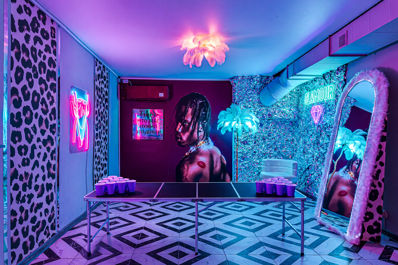 Арендовать Неоновый лофт Гламур. Квадратное помещение с чёрно-белым геометрическим полом. В центре — стол для пинг-понга с розовыми пластиковыми стаканами. Стены украшены неоновыми вывесками, постерами и зеркалом в пухлой раме. Освещение фиолетового оттенка, на потолке люстра.