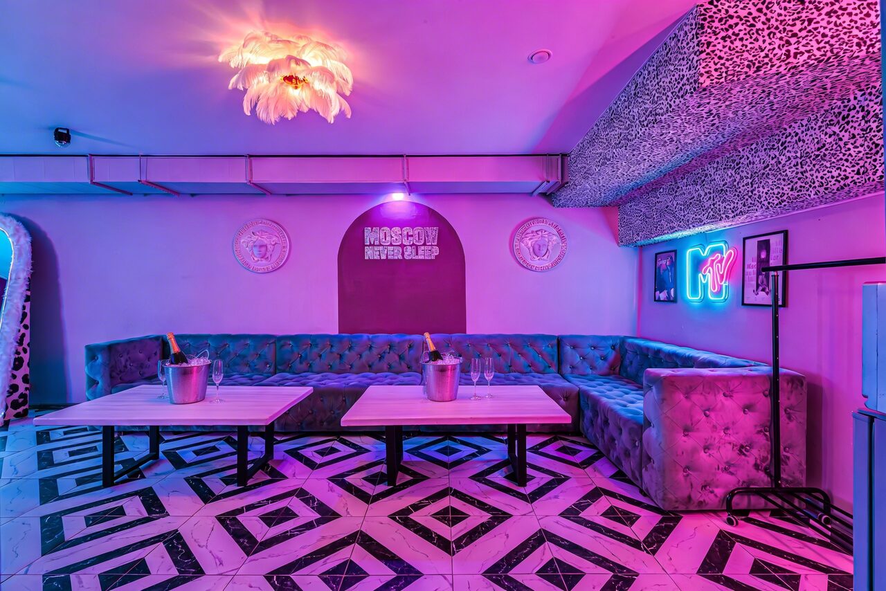 Арендовать Неоновый лофт Гламур. Клубное пространство с фиолетовым освещением, размещены мягкие угловые диваны серого цвета, два низких прямоугольных стола. Пол выложен плиткой с геометрическим рисунком. На стене неоновая вывеска, вокруг декор с леопардовым узором.