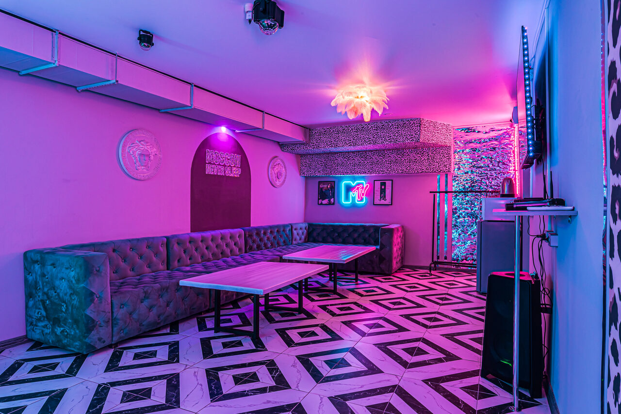 Арендовать Неоновый лофт Гламур. Пространство с мягкими диванами вдоль стен, два стола. Пол с геометрическим узором. Неоновое освещение, фиолетовые и розовые тона. На стенах декоративные элементы, телевизор, неоновая вывеска. Подвесной светильник, звукозаписывающее оборудование.
