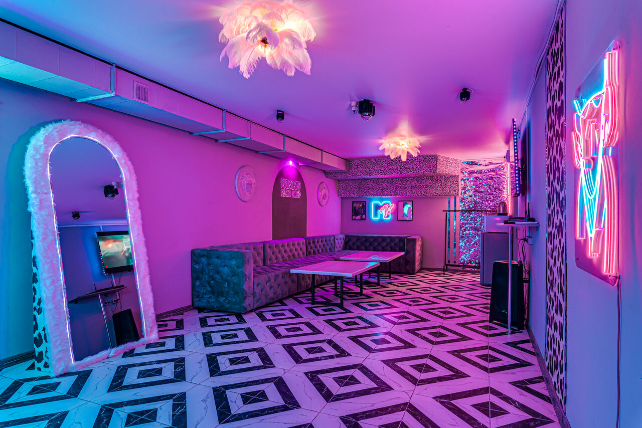 Арендовать Неоновый лофт Гламур. Пространство с неоновым освещением, мягкими диванами вдоль стены, прямоугольным столом, декоративными элементами на стенах, зеркалом с меховой рамкой и барной стойкой. Черно-белый геометрический узор на полу, розовые и синие неоновые вывески.
