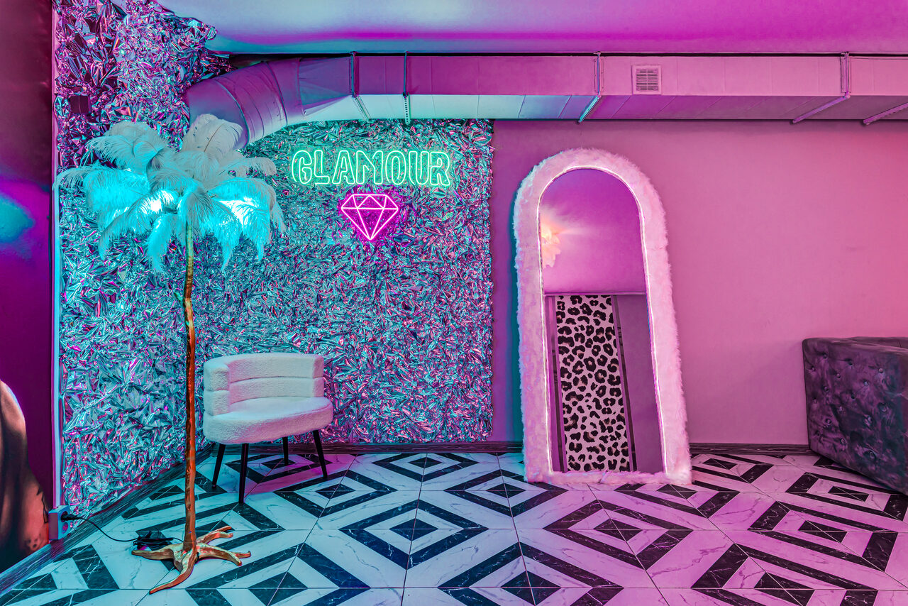 Арендовать Неоновый лофт Гламур. Пространство с неоновым словом "GLAMOUR" и изображением бриллианта на стене с узором, белое кресло, искусственная пальма, зеркало с меховой рамой, красочная плитка на полу. Освещение разноцветное.